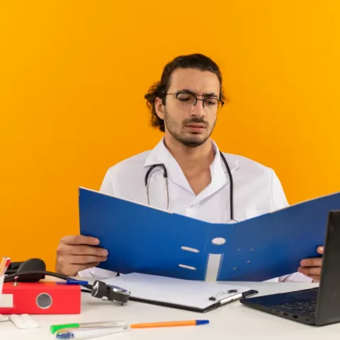 cunfused-young-male-doctor-with-medical-glasses-wearing-medical-robe-with-stethoscope-sitting-desk