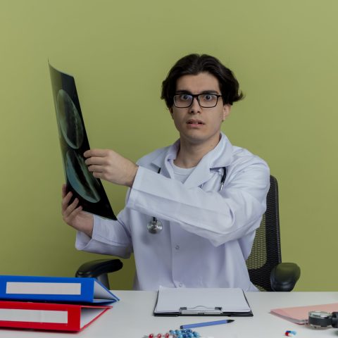 impressed young male doctor wearing medical robe and stethoscope with glasses sitting at desk with medical tools holding x-ray shot looking at camera isolated on olive green background