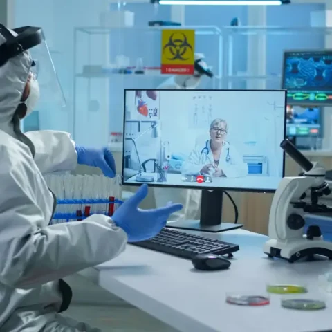 lab-technician-ppe-suit-sitting-table-medical-research-laboratory-holding-test-tube-talking-video-call-with-senior-doctor-giving-online-advices-virtual-meeting