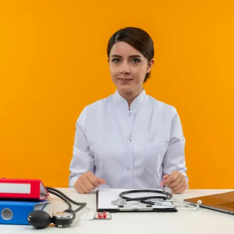 pleased-young-female-doctor-wearing-medical-robe-with-stethoscope-sitting-desk-work-computer-with-medical-tools-isolation-yellow-wall