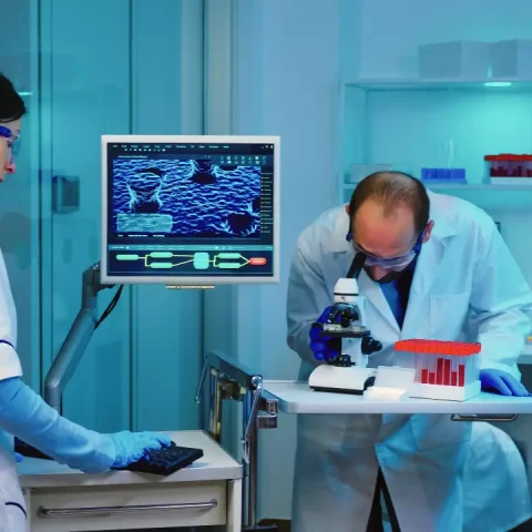 scientist-researcher-using-microscope-laboratory-while-nurse-taking-notes-computer-working-overtime