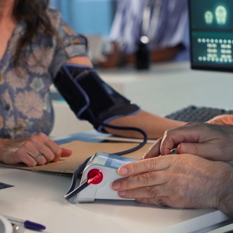Senior physician measuring blood pressure with a blood pressure monitor, using professional tools at clinical consultation. Old doctor does a tension reading and heartbeat measurement. Camera B.