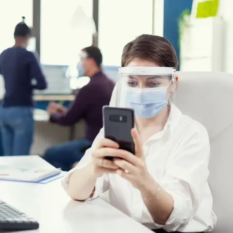 woman-with-protective-mask-working-professional-workspace-typing-mobile-phone-front-computer