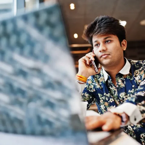 young-male-indian-freelancer-sitting-open-laptop-computer-fast-food-cafe-handsome-asian-man-speaking-phone-work-netbook-comfortable-coffee-shop